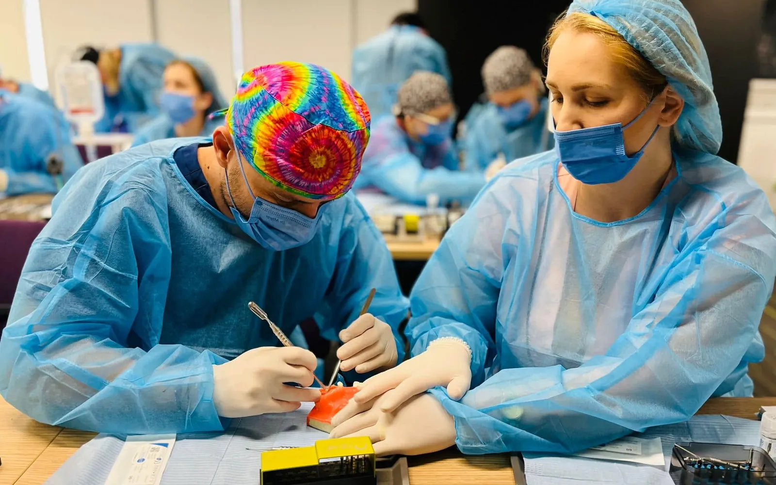 Dr. Ramona Drilea, în halat albastru de unică folosință, lucrează pe un model alături de un coleg, într-o sală de curs plină de medici.
