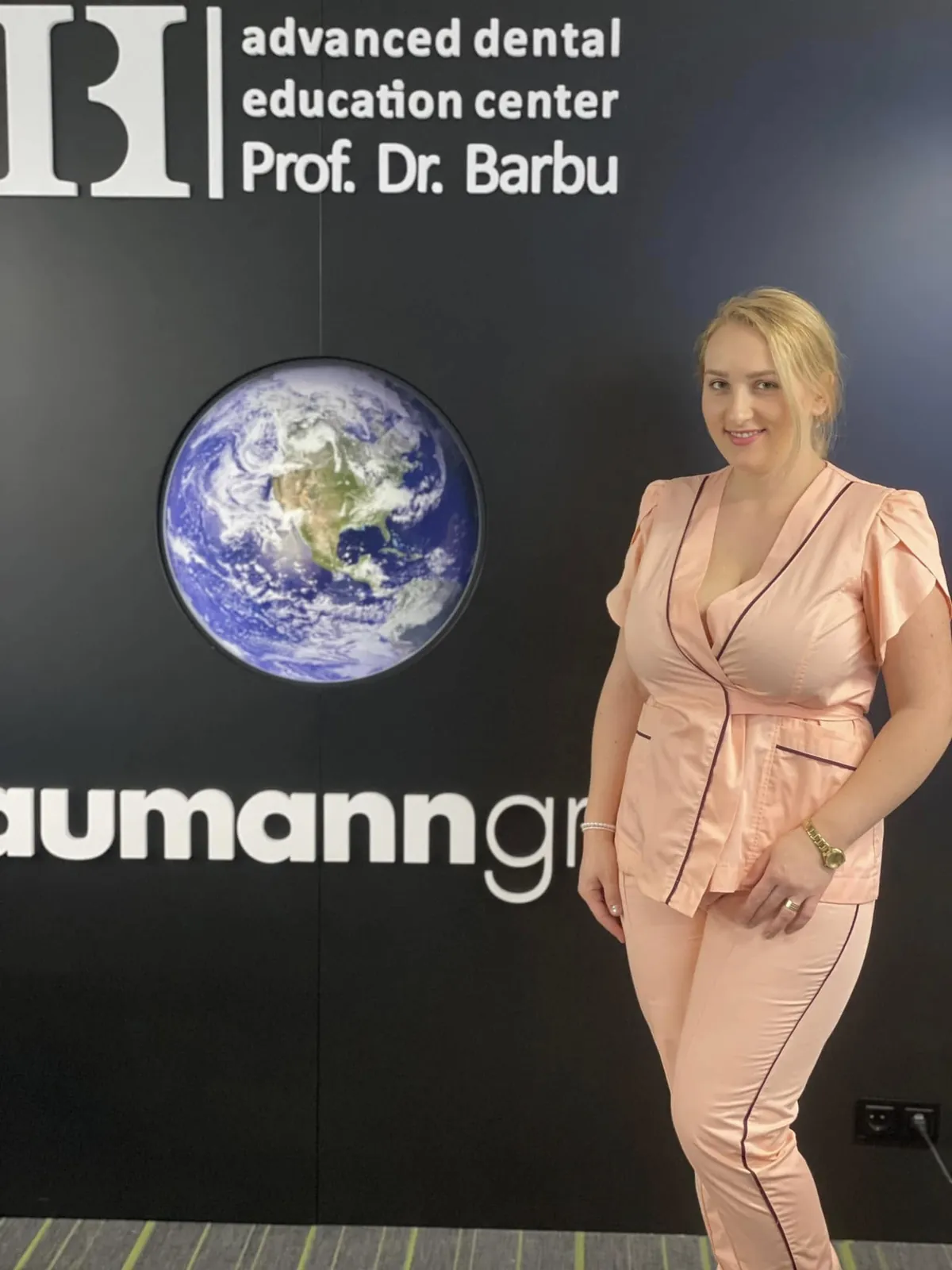 Dr. Ramona Drilea zâmbind, într-un costum medical roz, în fața peretelui advanced dental education center Prof. Dr. Barbu.